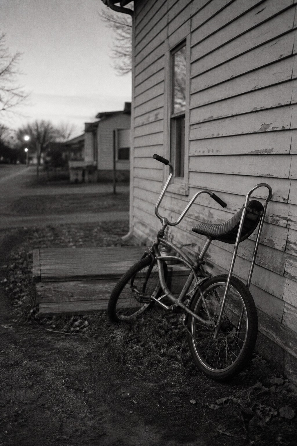 A child's bicycle from the 1970s leaning alone against the clapboard wall of a small house at dusk.