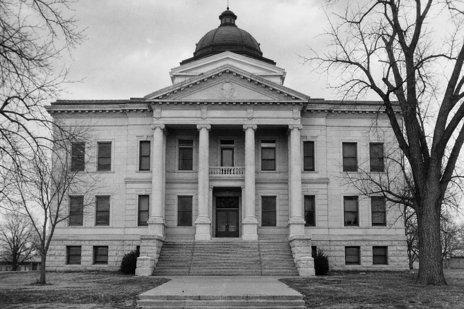 An austere county courthouse in winter, bare trees in front of stone steps under flat, overcast light.