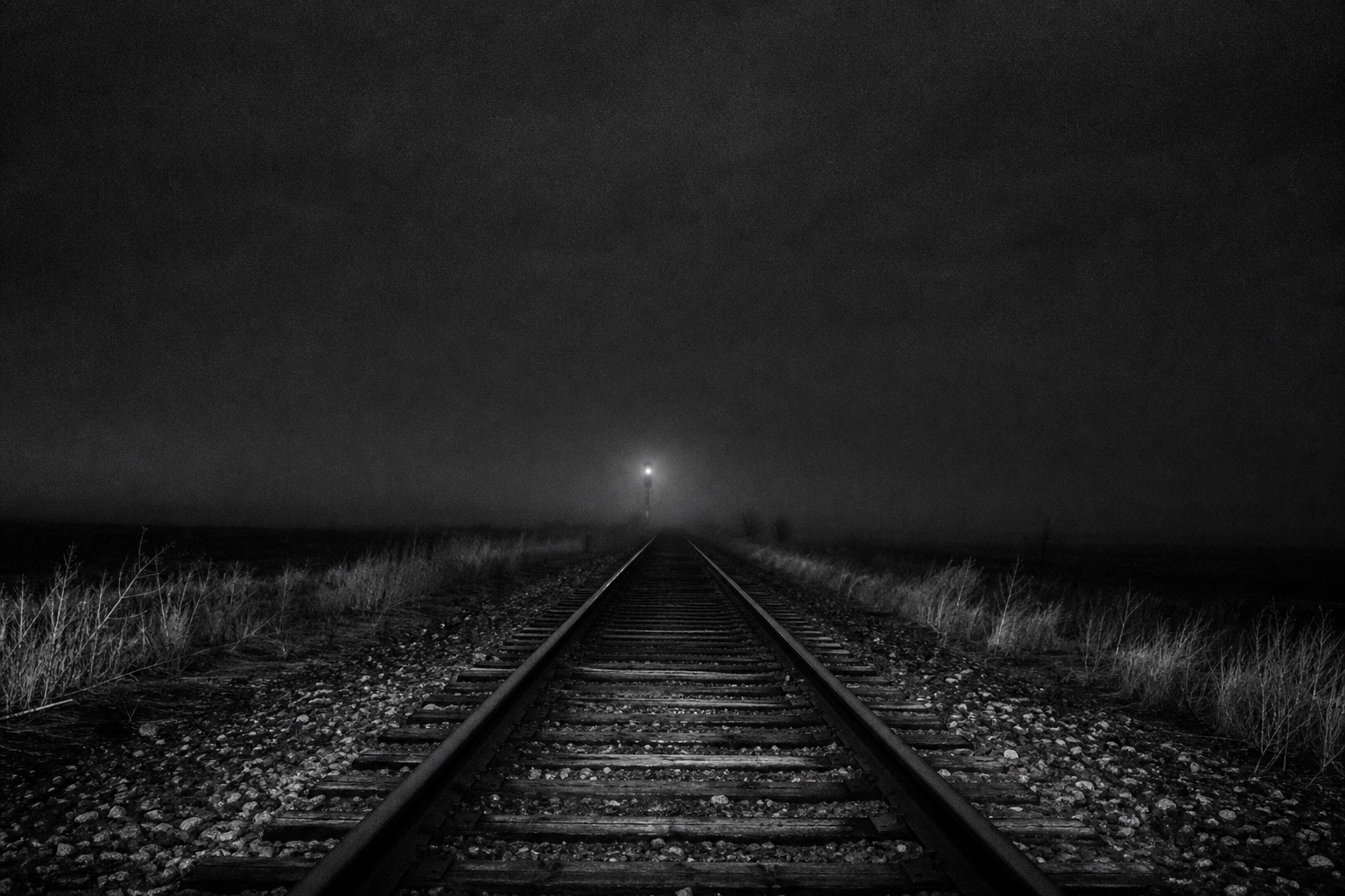 Railroad tracks receding into darkness and low fog at night, a faint signal light in the far distance.