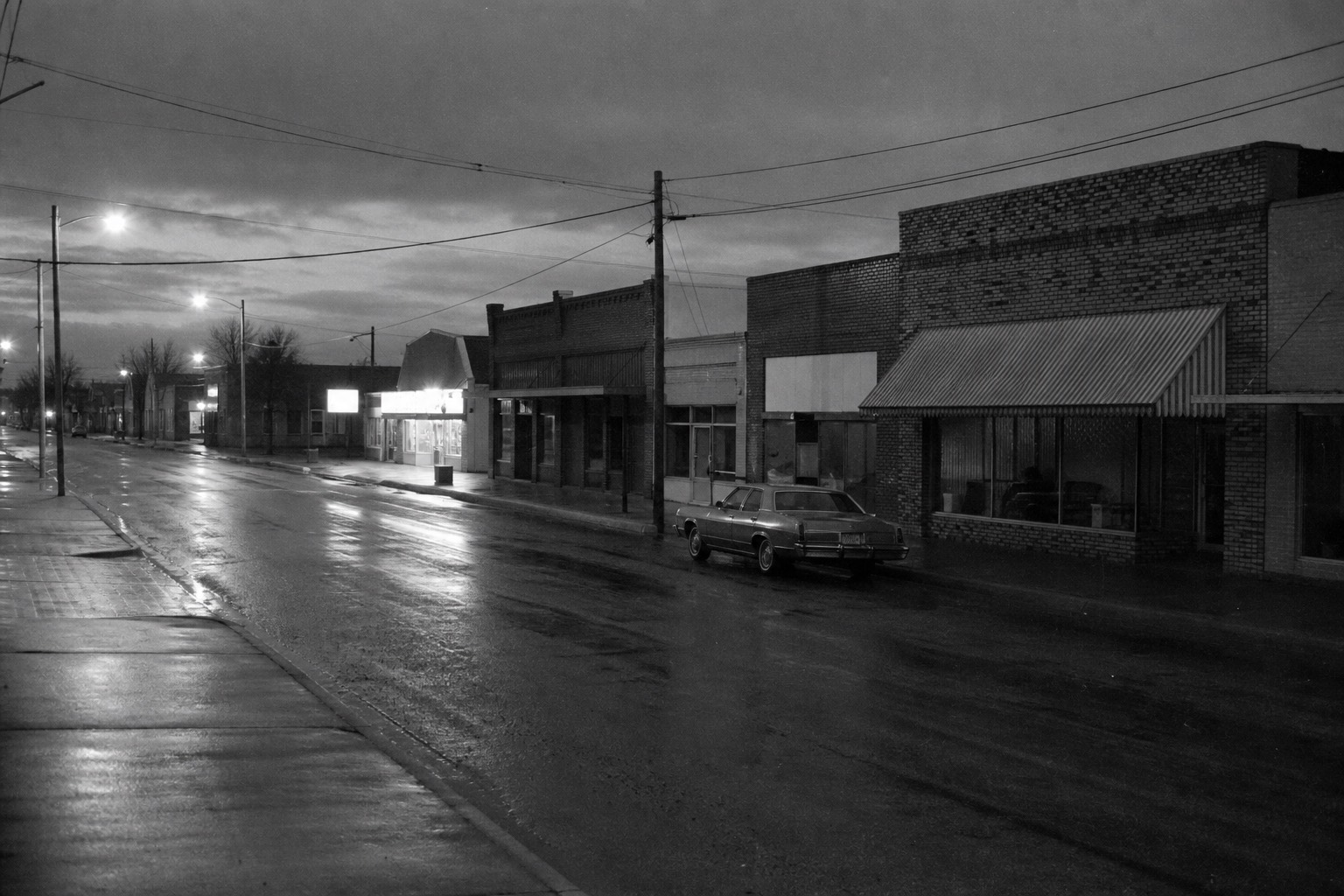 A small-town Oklahoma main street on a cold evening in the late 1970s, storefronts dark, one distant sign lit.