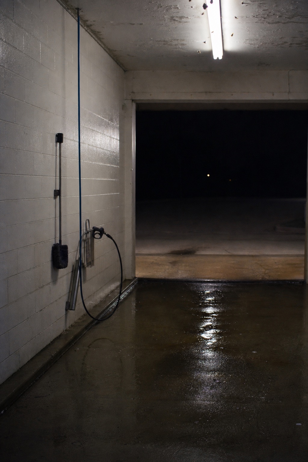 The interior of an empty car wash bay at night, a spray wand hanging on the wall beneath a bare fluorescent tube.