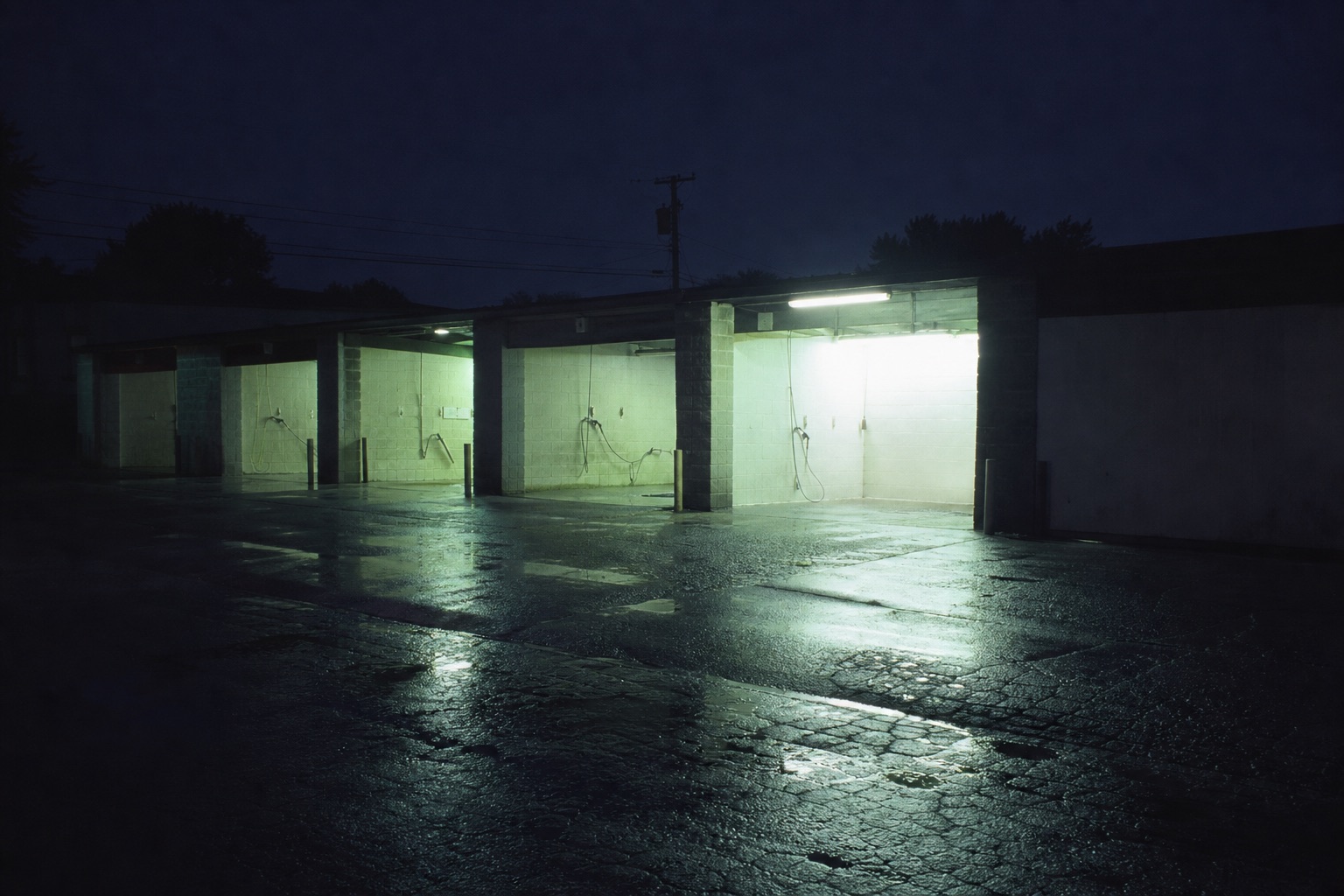 A self-service car wash at night, cold fluorescent light spilling from the open bays onto wet asphalt.