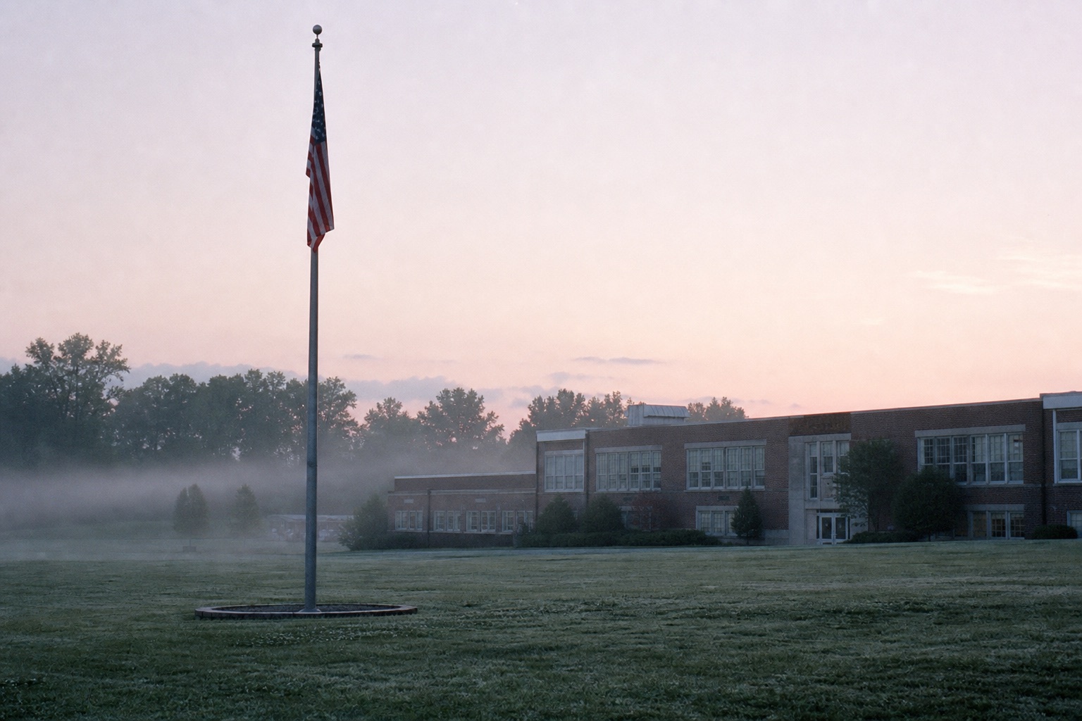 A suburban high school campus at dawn, a flagpole on a dewy lawn in thin morning fog.
