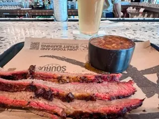Sliced smoked brisket on a paper-lined tray with baked beans and a pale lager at the 4 Rivers airport counter