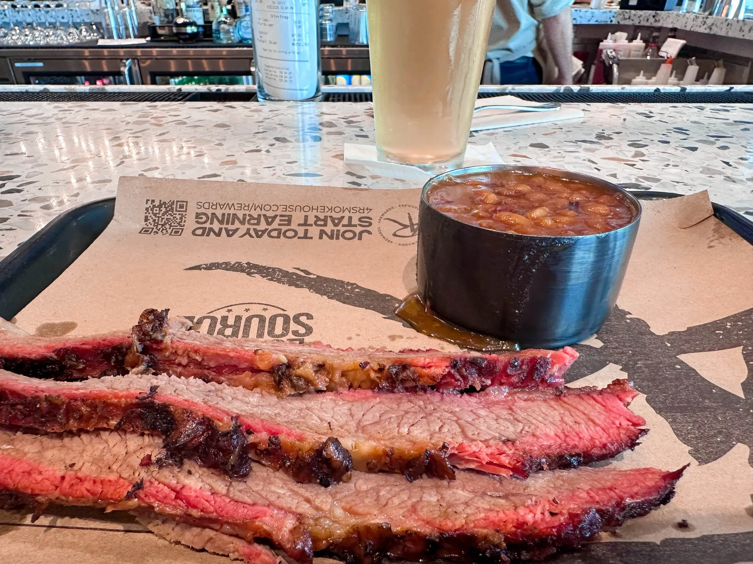 Sliced smoked brisket on a paper-lined tray with baked beans and a pale lager at the 4 Rivers airport counter