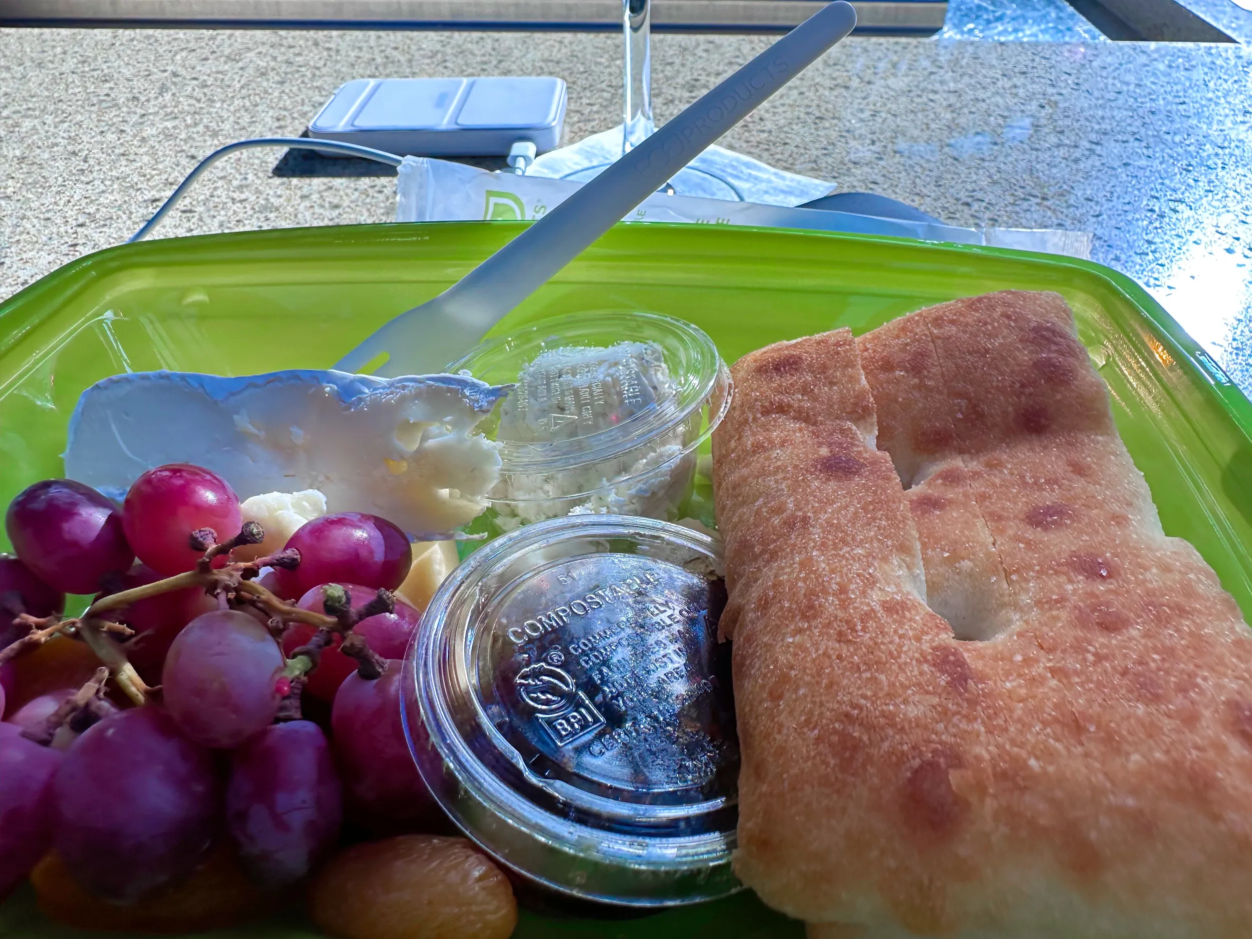 Cheese plate at Beaudevin in O'Hare airport: soft cheese, red grapes, dried apricots, focaccia, and compostable cups of spread in a green takeout container, with a wine glass and phone charger on the granite counter behind.