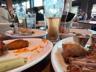 Shared plates of smoked wings and halved brisket Reuben egg rolls with dipping sauces, celery, water glasses, and a pale draft beer on a crowded restaurant table