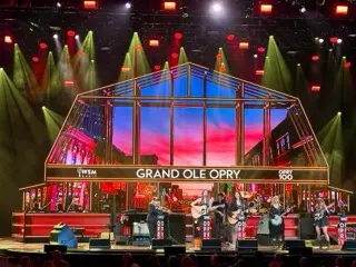 Grand Ole Opry stage with glowing house-shaped set, GRAND OLE OPRY sign, WSM Radio and Opry 100 branding, green spotlights, and multiple performers onstage