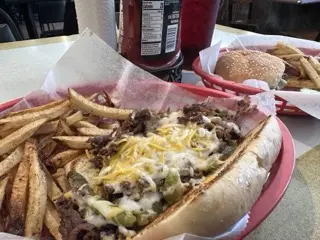 Two red baskets on a restaurant table: an open Philly sandwich with steak, peppers, onions, and melted cheese beside fries in front, with a sesame-seed burger, fries, and two Coke bottles behind