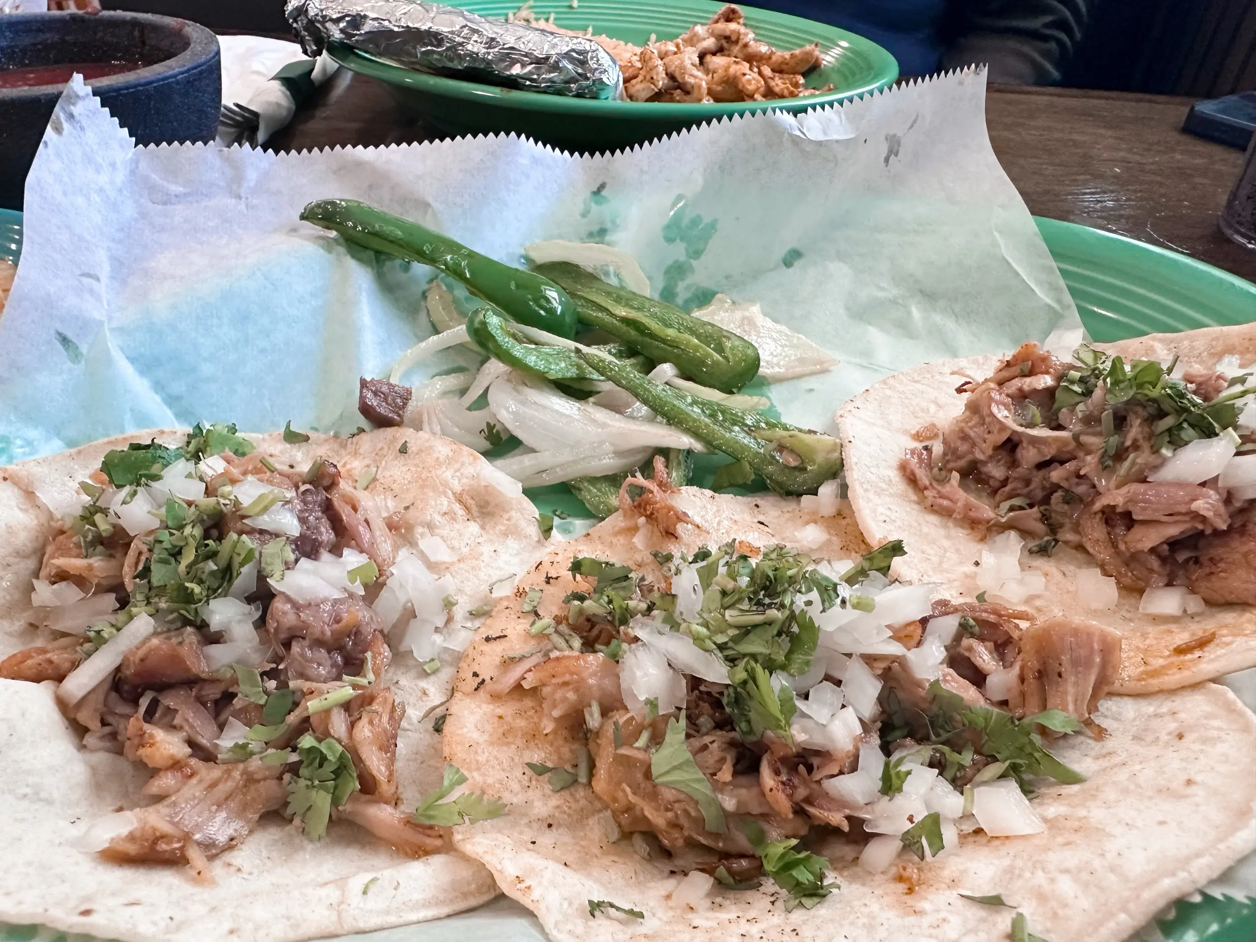 Close-up of carnitas street tacos topped with chopped onion and cilantro beside grilled jalapeños and onions, with arroz con pollo in the background