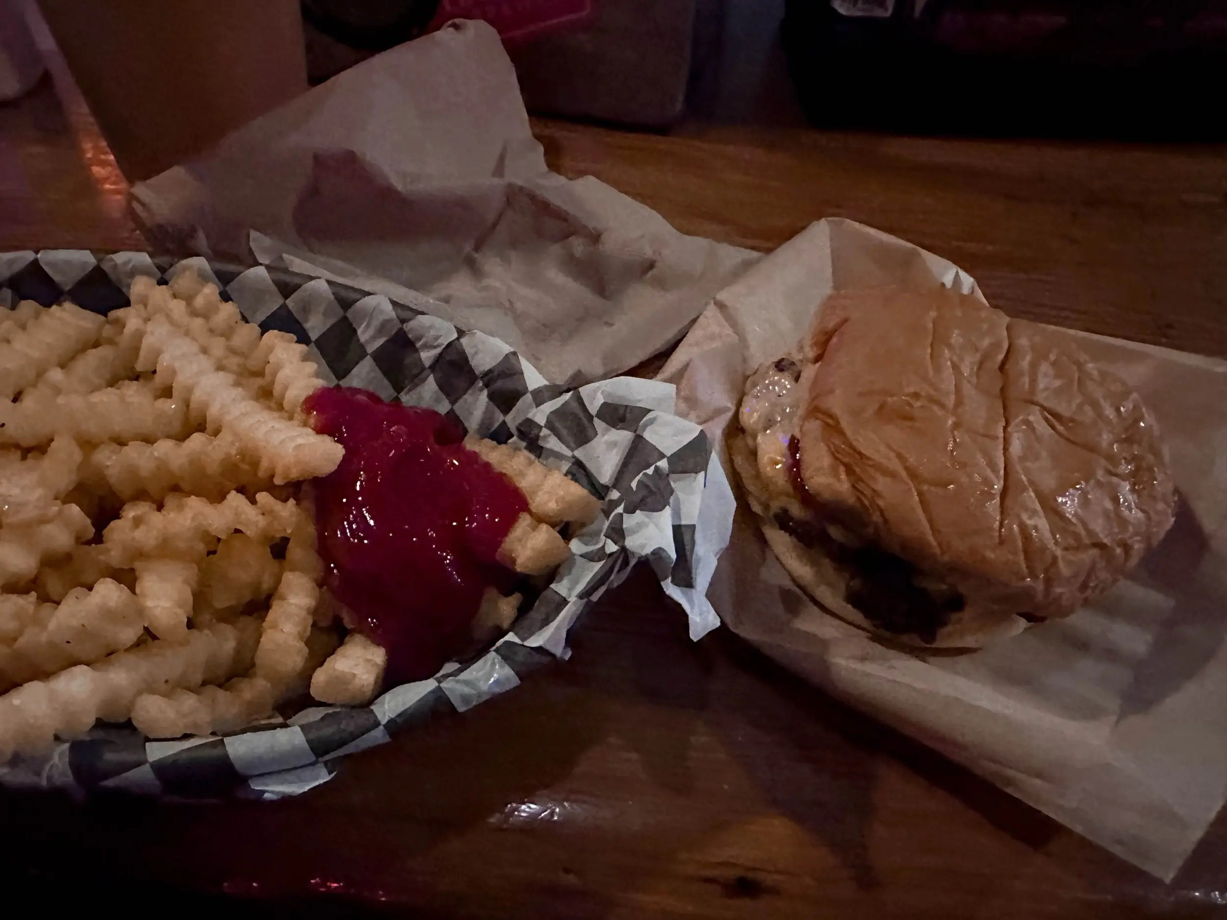 A burger on a soft bun beside crinkle-cut fries with ketchup at Jack Brown's Columbia, with a pale draft beer just visible in the dim bar light