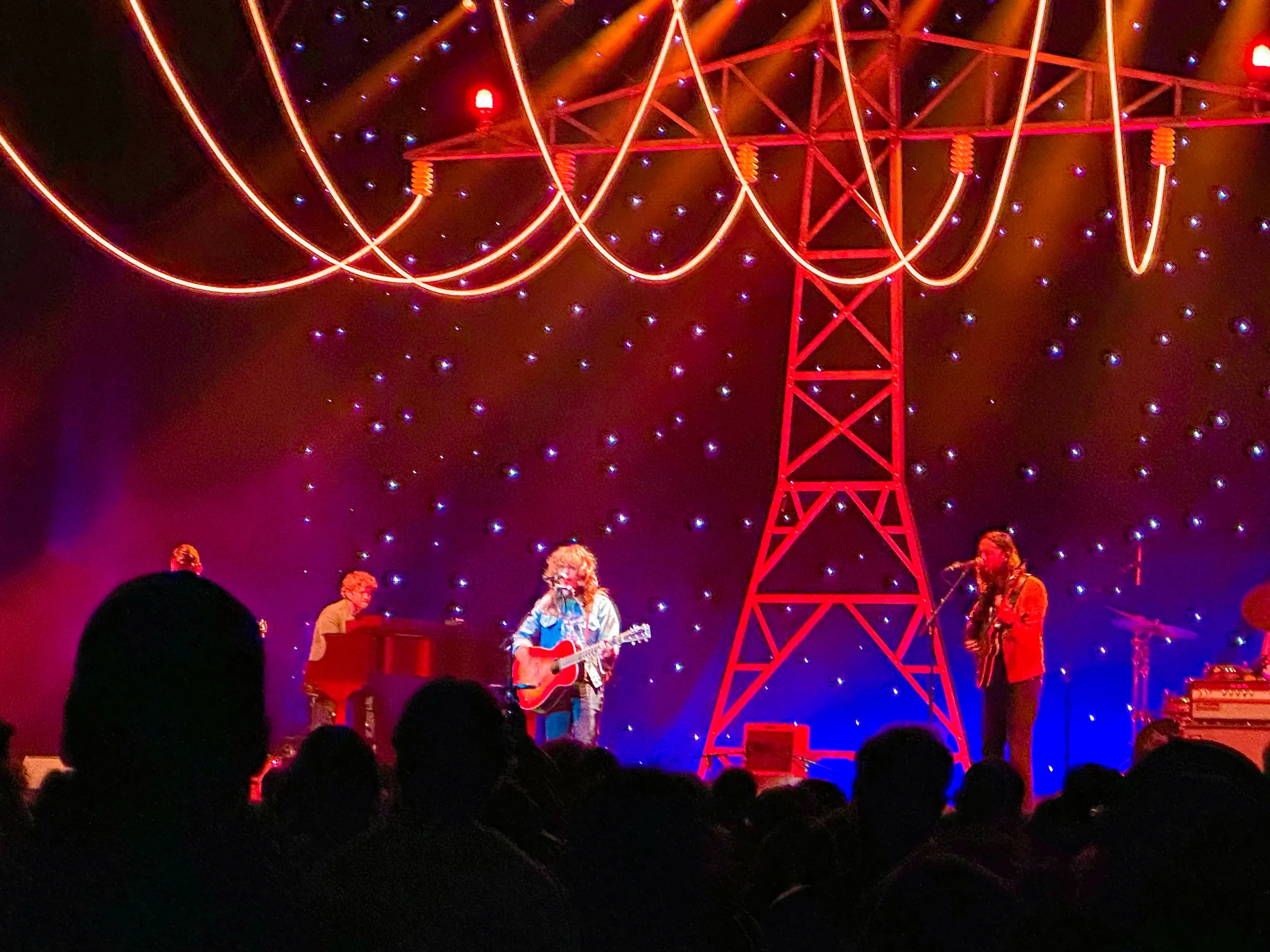 Jesse Welles performs with an acoustic guitar at The Pinnacle, flanked by a pianist and a guitarist, beneath a glowing red electricity pylon and a starry backdrop as silhouetted fans watch.