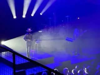 LP performing with a guitar and broad-brimmed hat beneath white spotlights cutting through deep blue stage haze at Brooklyn Bowl Nashville