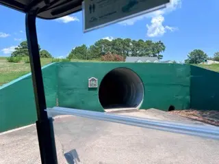 View from a golf cart at Myrtlewood Golf Club facing a wide green tunnel under a path, with bright summer sky, trees, and low buildings beyond the embankment.