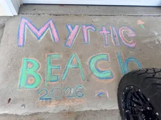 Colorful chalk writing on concrete that says 'Myrtle Beach 2026,' with part of a large golf car tire visible at the right edge