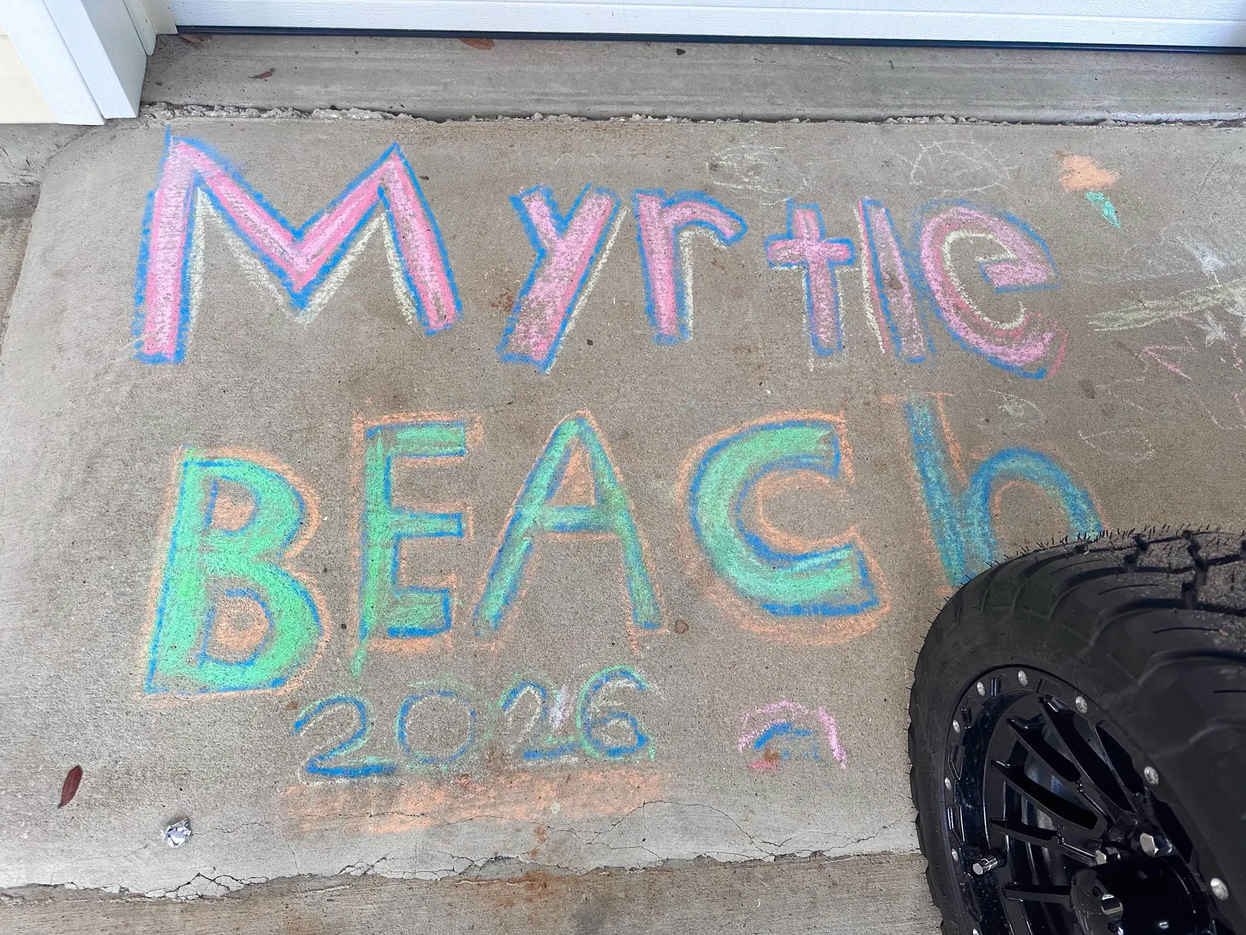 Colorful chalk writing on concrete that says 'Myrtle Beach 2026,' with part of a large golf car tire visible at the right edge