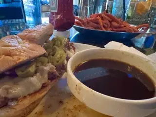 Close-up of a sandwich with melted cheese and pickled jalapeños, a bowl of dark dipping sauce, and a plate of sweet potato fries on a table.