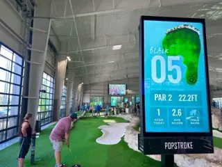 Indoor PopStroke putting course with two players beside the green and a large digital sign showing the Black course hole 05, par 2, 22.2 feet.