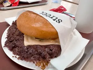 A cheeseburger at Street Smash Burgers in Matosinhos with a glossy bun, a dark lacy-edged smashed patty, melted cheese, branded wrapper, and a white drink cup on a cafeteria tray
