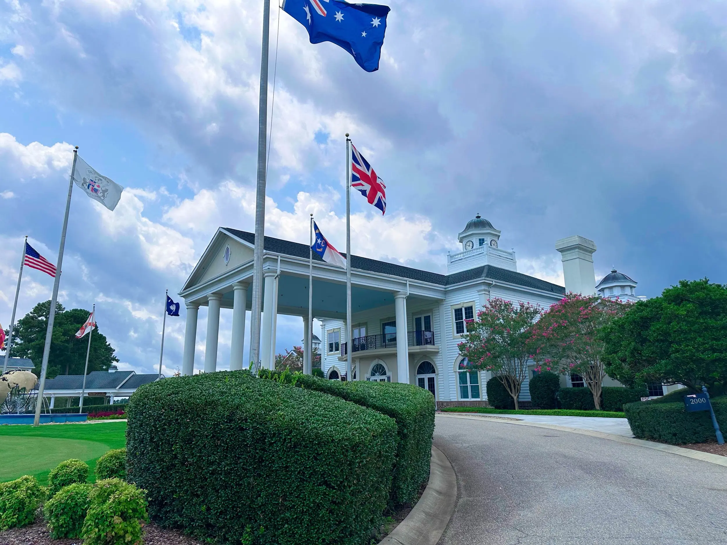 White columned clubhouse at World Tour Golf Links with trimmed hedges, a curved driveway, multiple national flags, and a dramatic blue-and-gray stormy sky above Myrtle Beach.